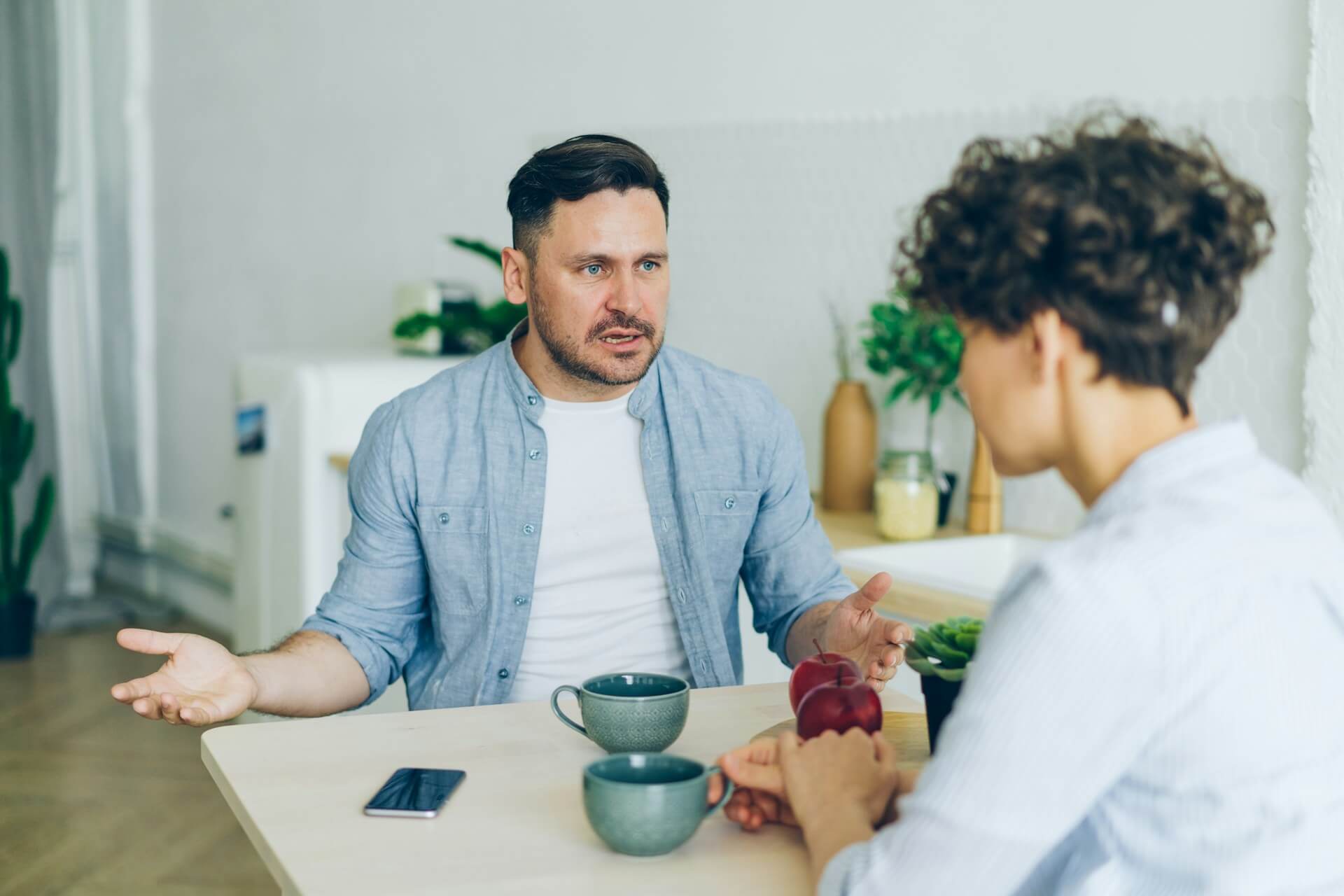 Man gesturing with open hands as he talks to a woman across a kitchen table with two mugs between them